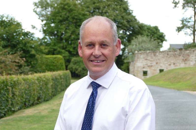 Man in white shirt and dark tie in front of a hedge and stone wall.