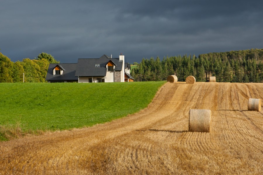 Bails of hay in a ploughed field next to grass with a house and woods in the background